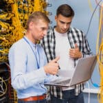 two men looking at a laptop in a server room